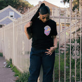Female model wearing black t-shirt with Oaklandish tree logo and heart graphic image on wearer's left chest standing in front of gate.