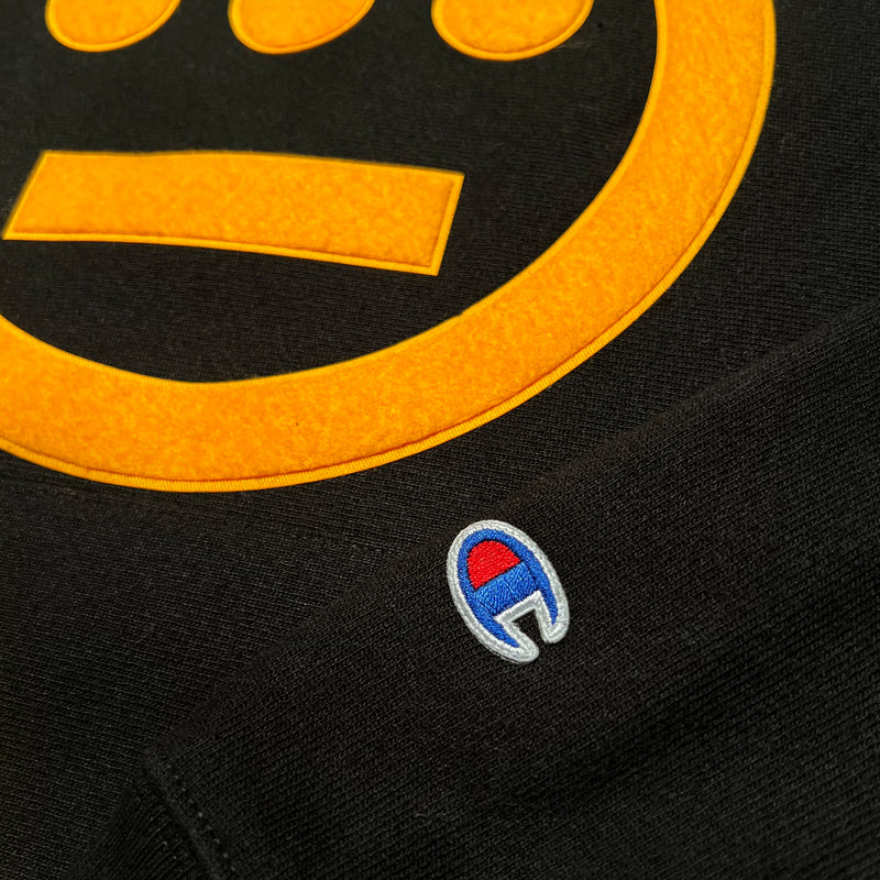 Detailed close-up of gold Hieroglyphics hip-hop logo on the chest and Champion logo on the sleeve of a black hoodie.
