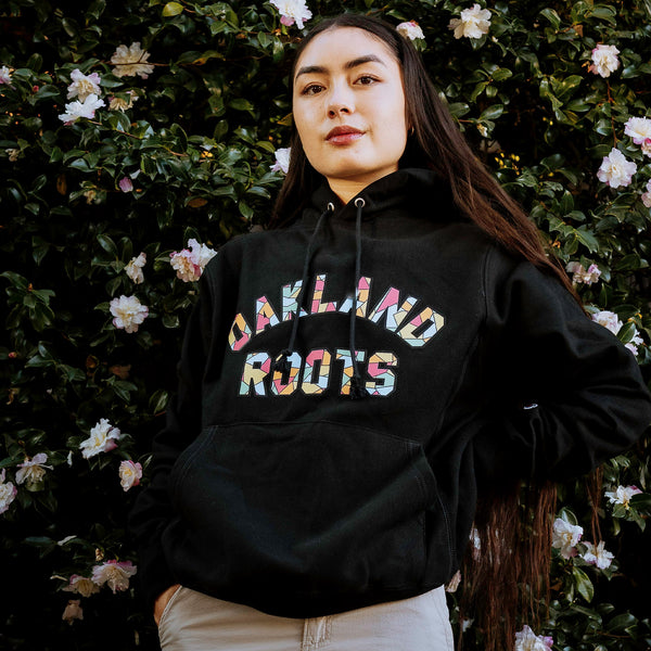 Female model wearing Champion reverse weave black pullover hoodie with Oakland Roots Mosaic pattern within applique wordmark 'Oakland Roots'.
