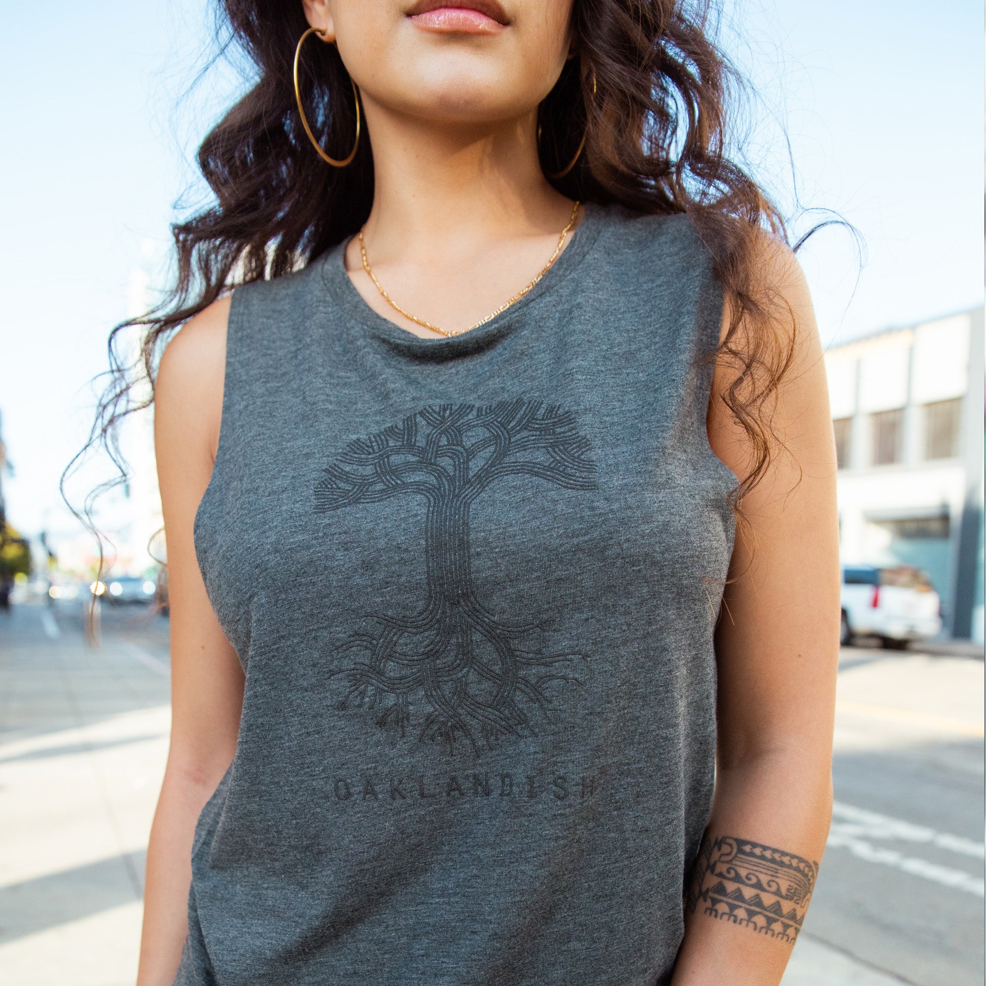 Detail close up of female model wearing Grey women’s cut tank top with black classic Oaklandish tree logo and wordmark on chest.