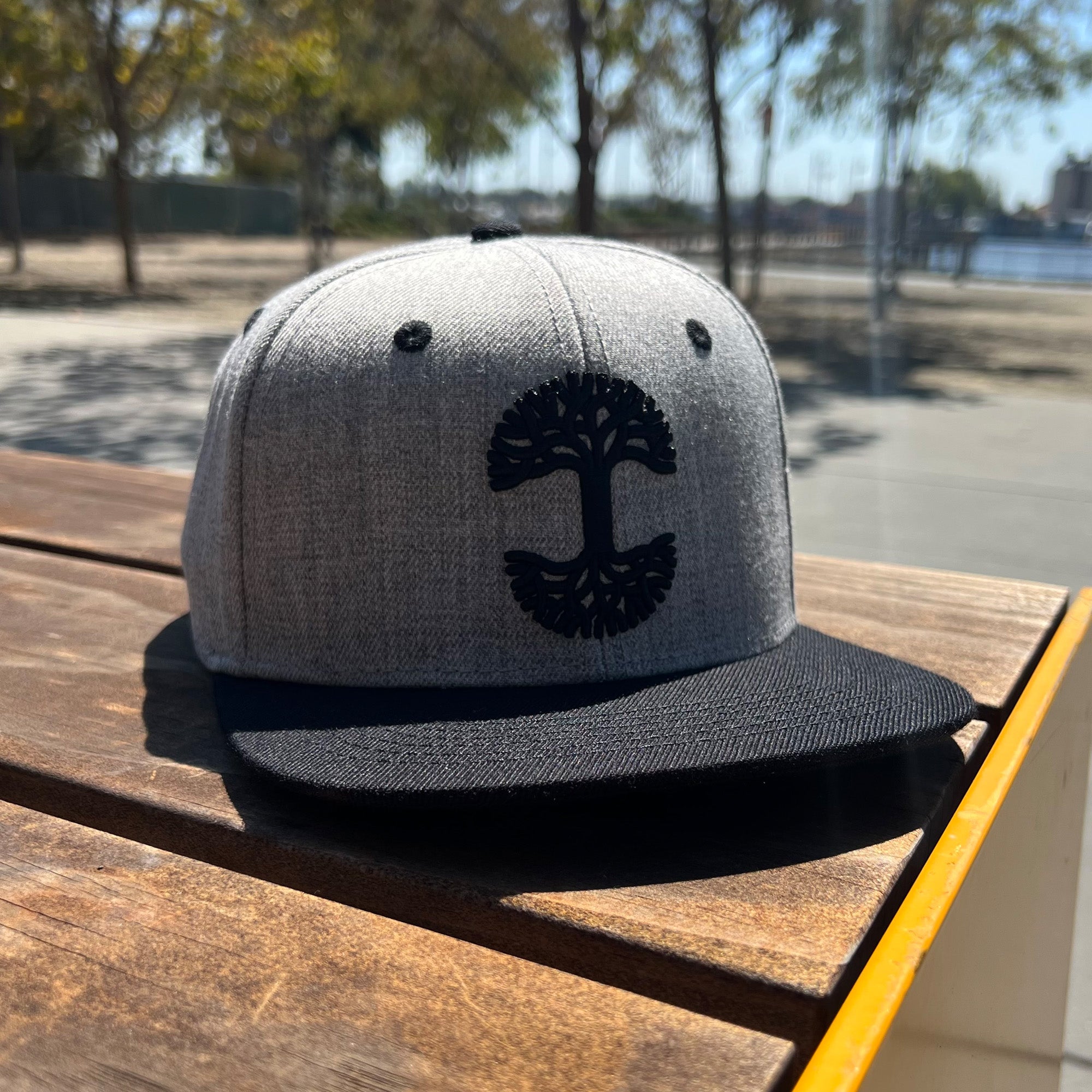 Photo image of Grey cap with black embroidered Oaklandish tree logo on the front outside in natural lighting on bench.