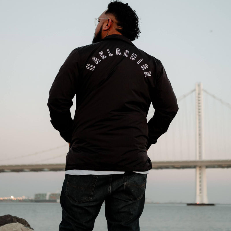 Back view of man on beach with Oakland bridge in background, wearing black coaches jacket with Oaklandish wordmark on the back. 