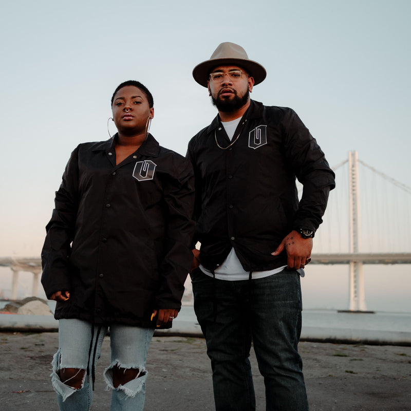Woman and man standing on beach with Oakland bridge in background wearing black Oaklandish coaches jacket.