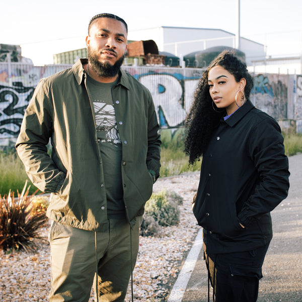 Man & woman in urban area with graffiti in background. Man a wearing green and woman wearing a black cotton, collared, snap close jacket.