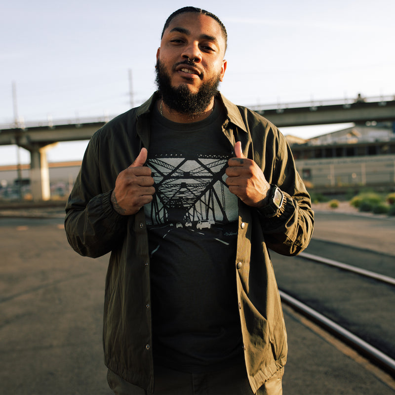 Man standing by rail road, wearing open snap close cotton jacket over Oaklandish t-shirt.