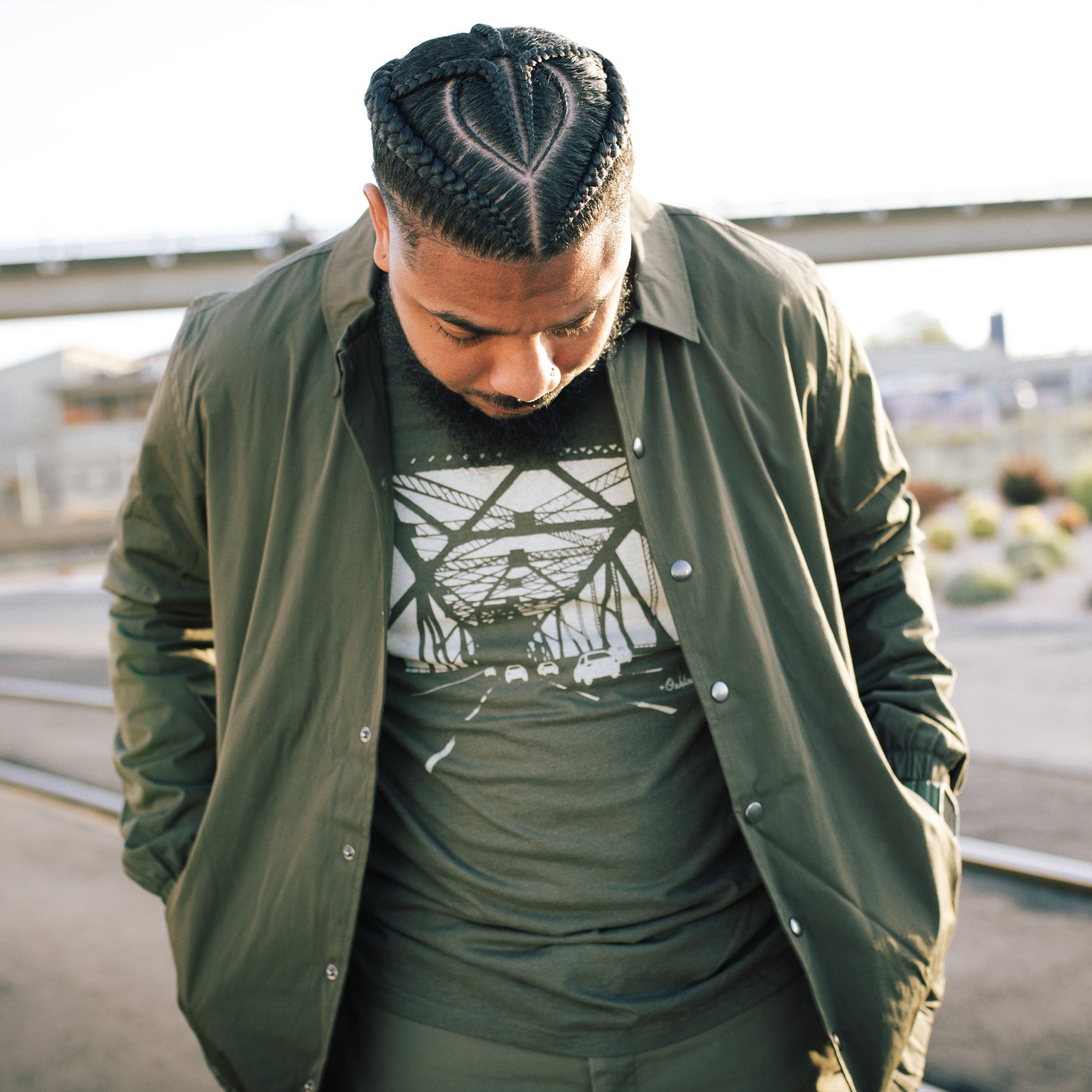 A man with neatly braided hair looks downward while wearing the Oaklandish Premium Coaches Jacket over a graphic t-shirt. He has his hands in his jacket pockets. The background features an industrial setting with a blurred view of a fence and some distant structures, giving the scene a casual and reflective mood.