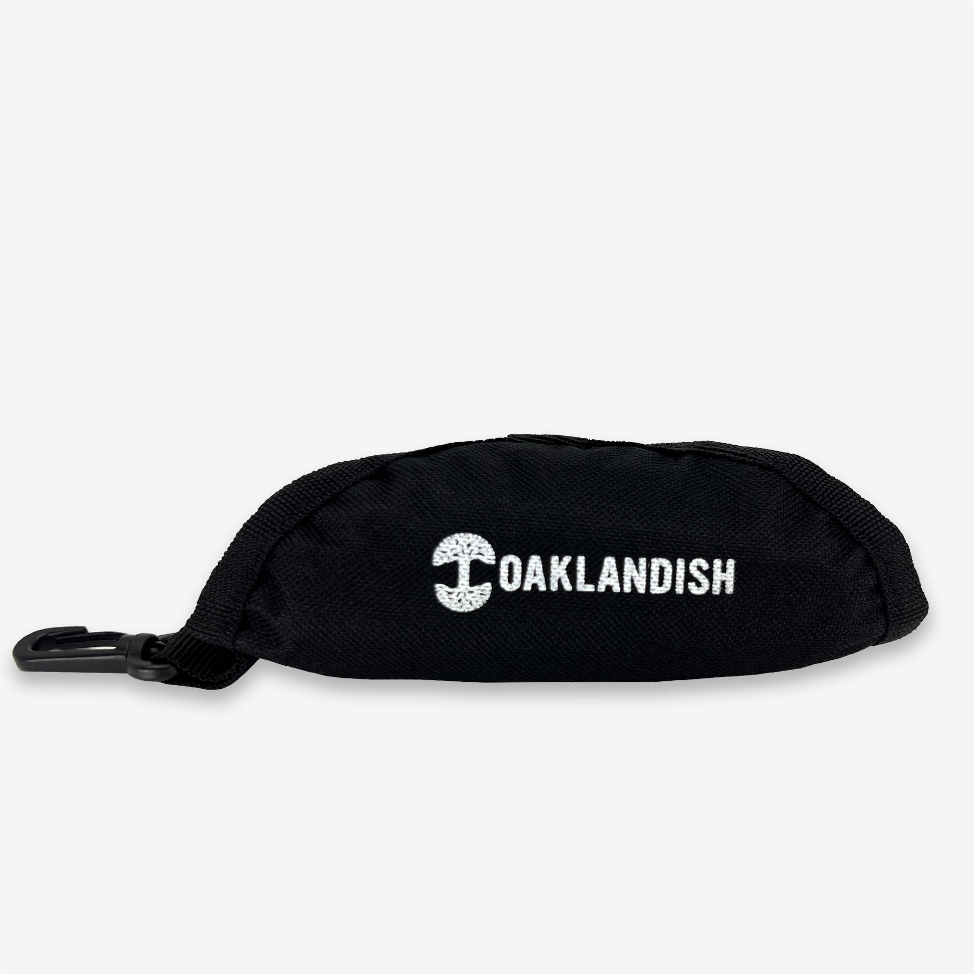 The Oaklandish Travel Pet Bowl is a small pouch made of durable black 600D polyester, featuring the word "OAKLANDISH" and a distinctive tree logo printed in white on the front. This versatile accessory includes a zipper and a plastic clip for easy fastening, making it perfect for carrying pet essentials. The background is plain white.
