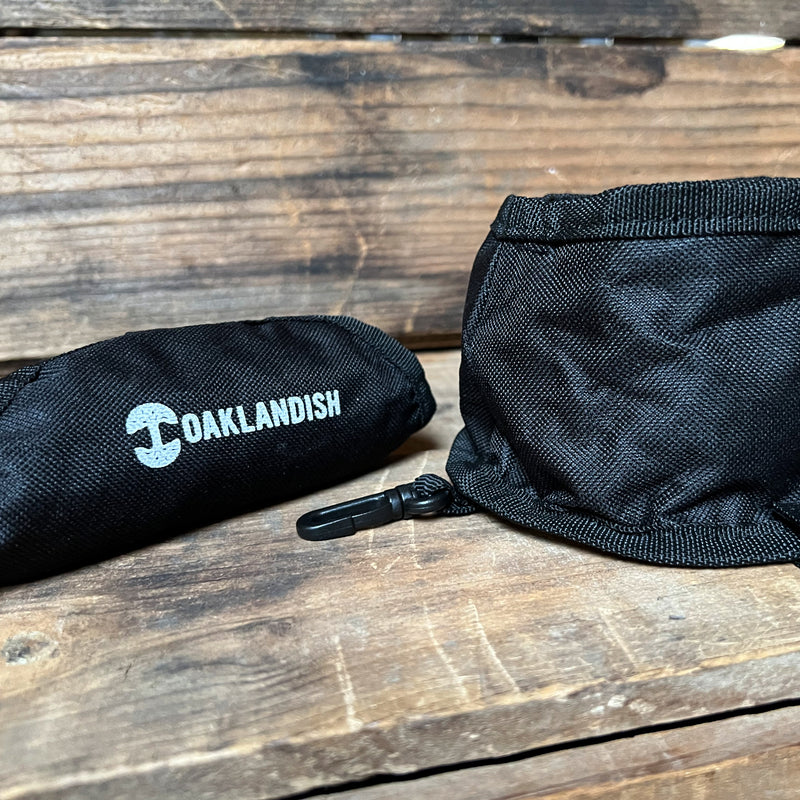 Black Oaklandish travel pet bowl and its stuff sack sitting outdoors on a wooden bench.