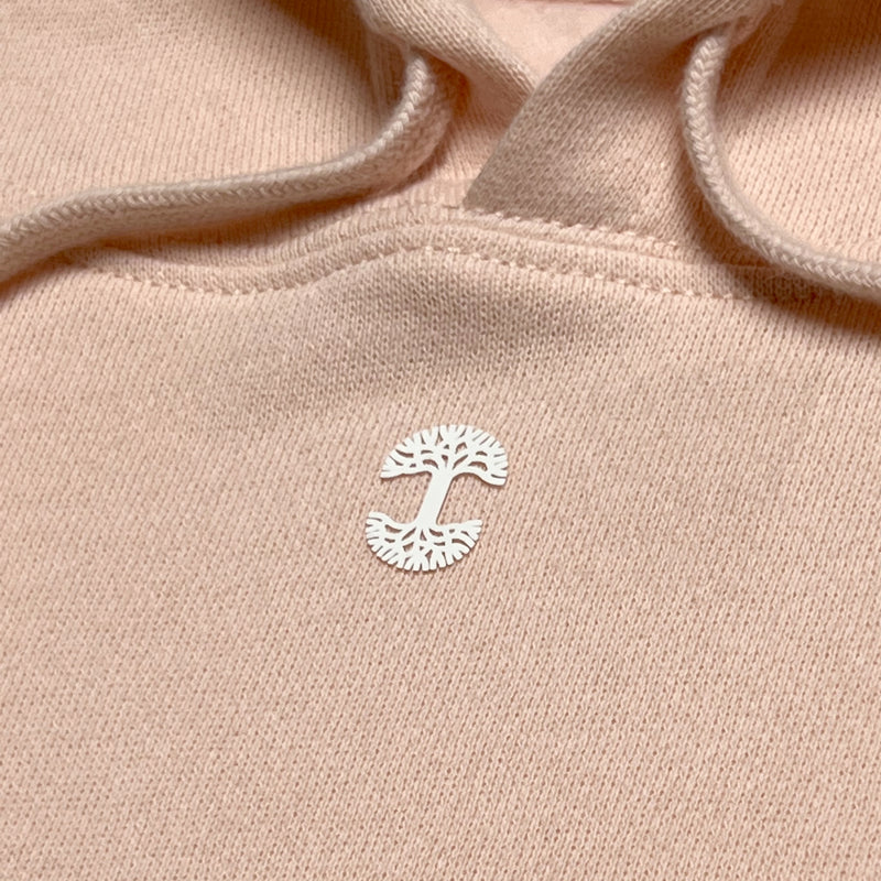 Close-up of white micro-sized Oaklandish tree logo on the chest of a short cropped women’s blush pink hoodie.