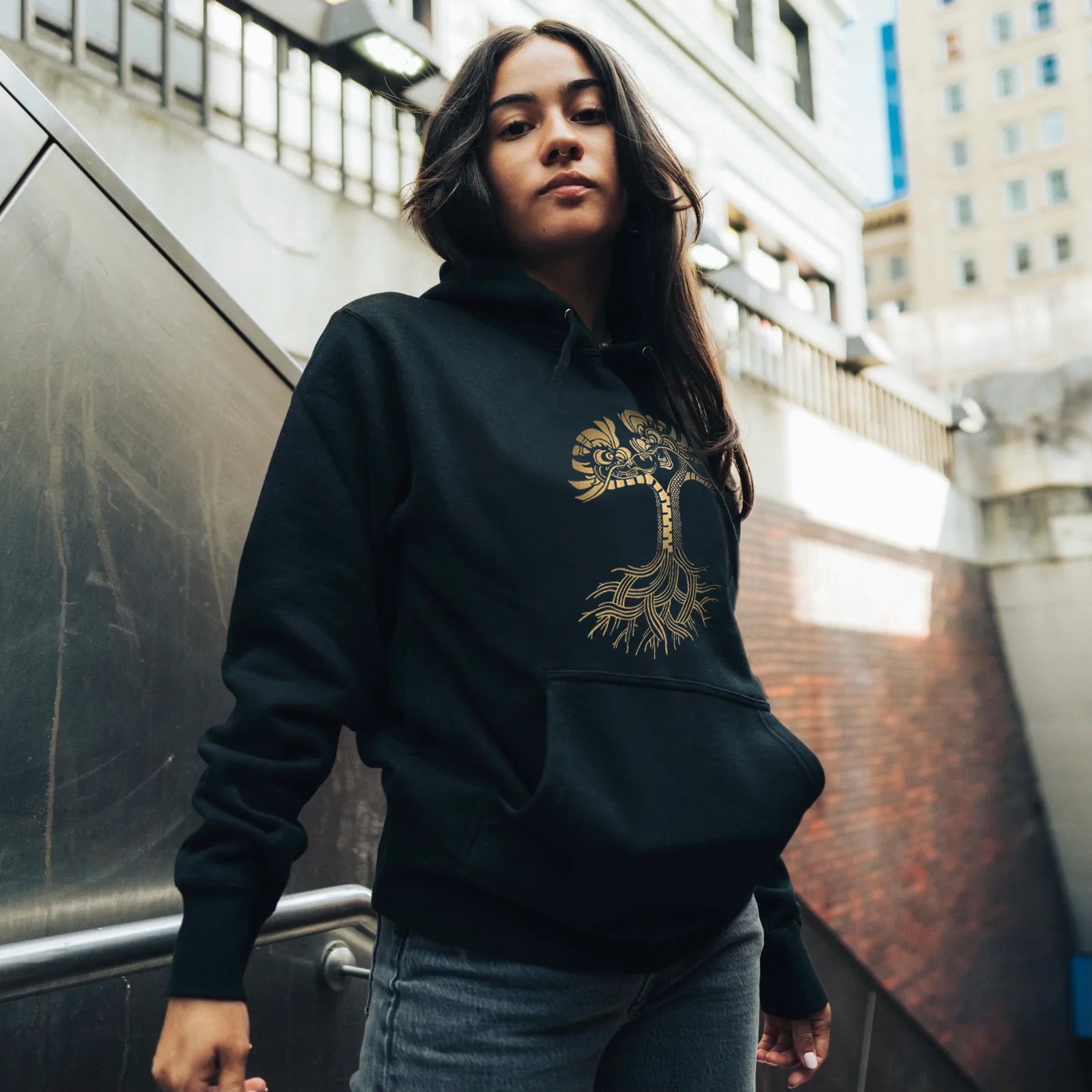 Female model outdoors in Oakland wearing black pullover hoodie with metallic gold dragon power design in the shape of the Oaklandish tree logo.
