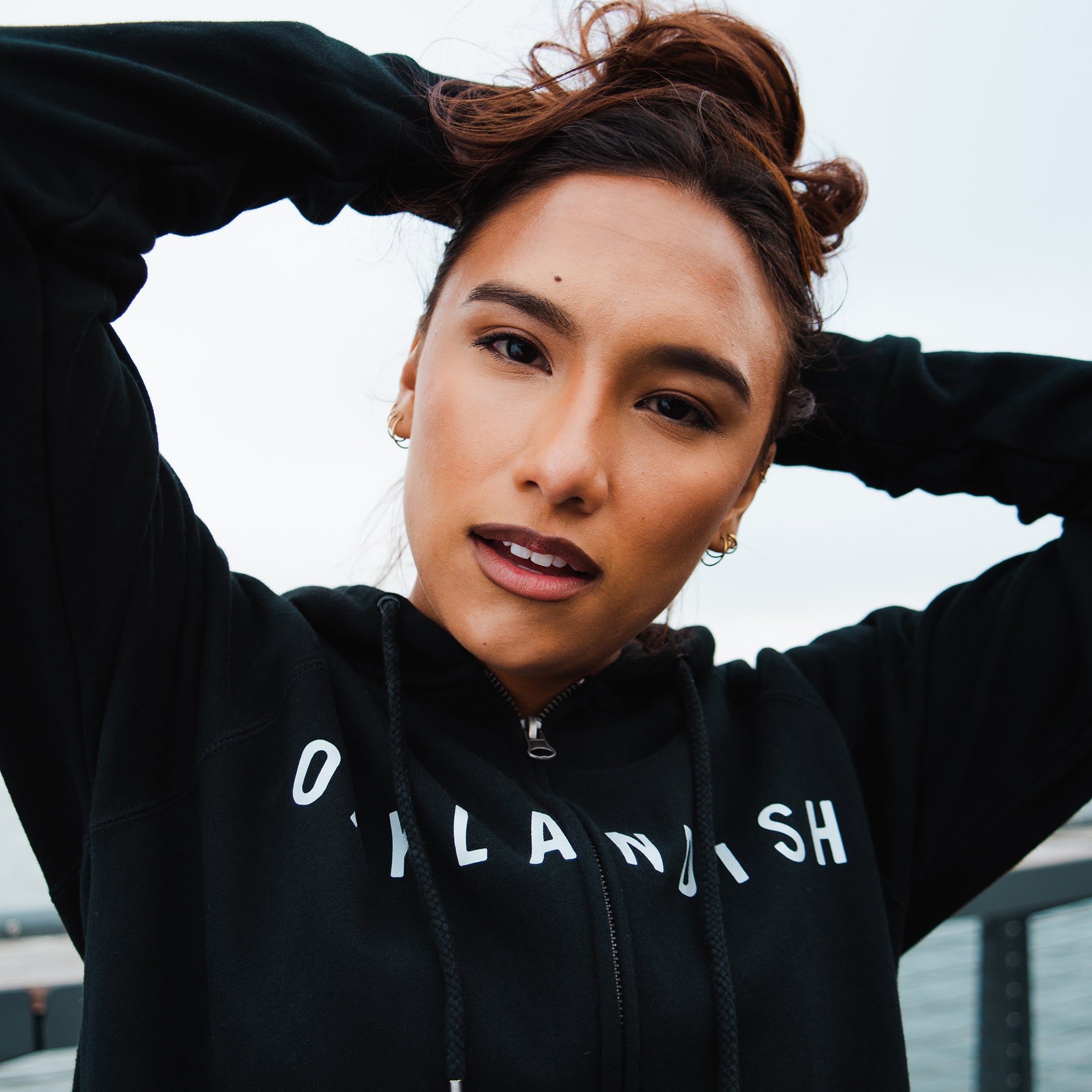 Close up of woman standing on a waterfront boardwalk wearing a black hooded sweatshirt with an Oaklandish wordmark on the chest.