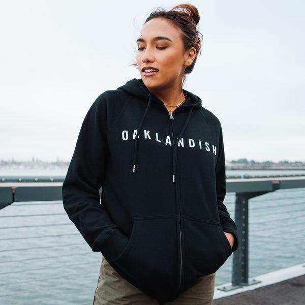 Woman standing on a waterfront boardwalk wearing a black hooded sweatshirt with an Oaklandish wordmark on the chest.