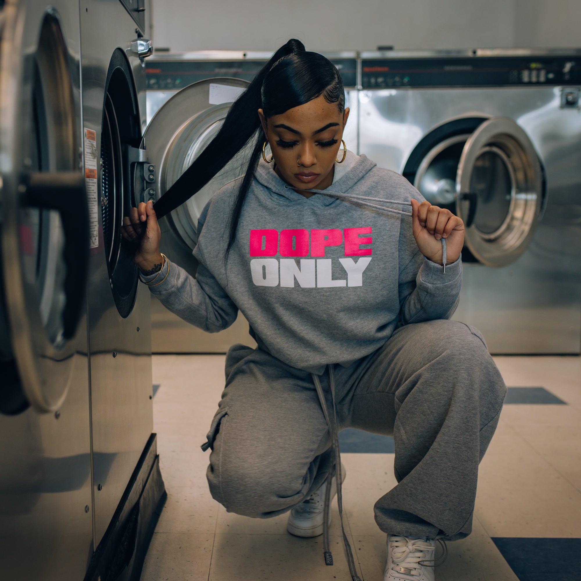 Female model at laundry mat wearing women's cropped athletic heather pullover hoodie with pink and white 'Dope Only' wordmark centered across the chest.