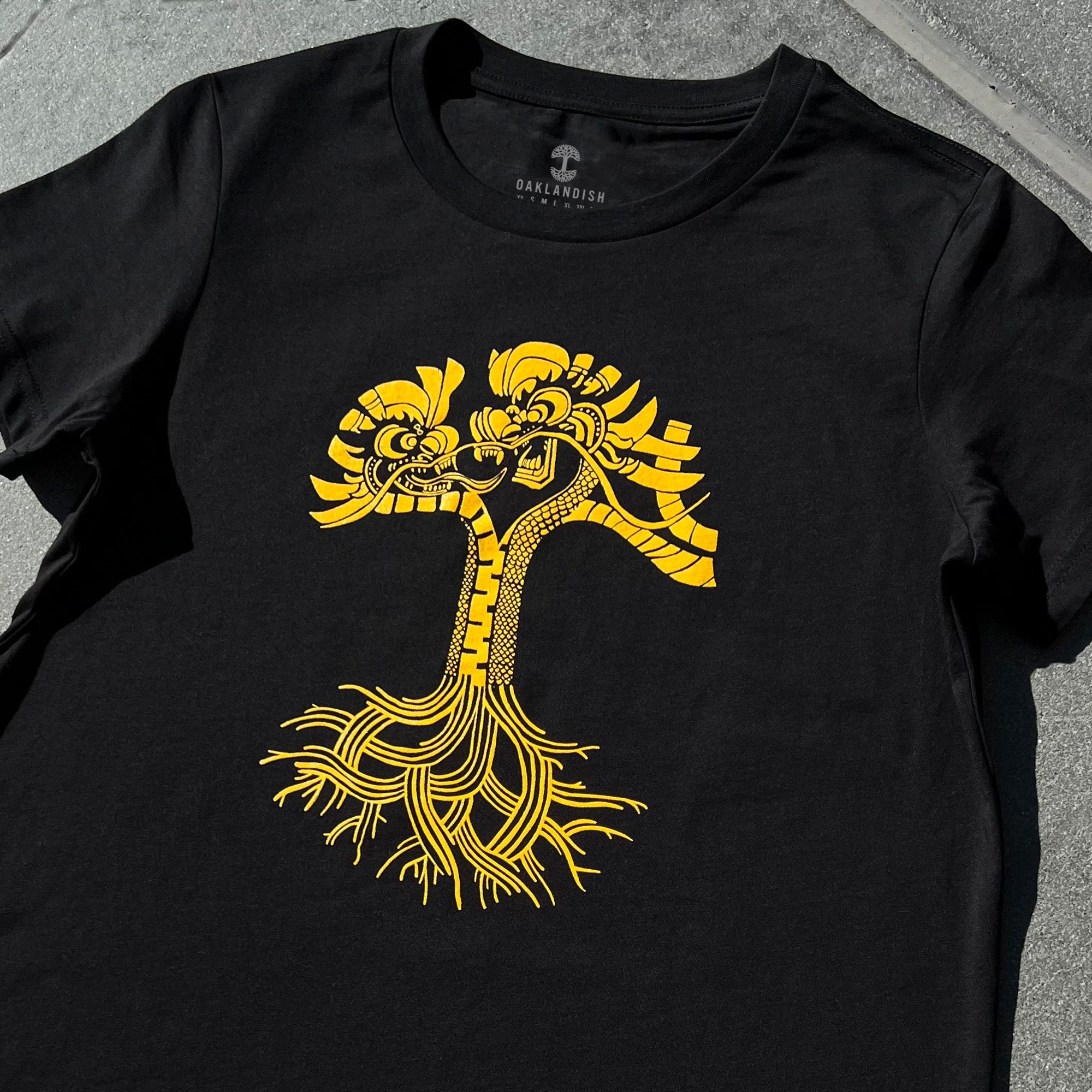 A black Women's Dragon Power Tee by Oaklandish, featuring a striking yellow graphic of an abstract, symmetrical design reminiscent of intertwined faces with root-like extensions. Laid flat on a gray surface, it prominently displays the "Oaklandish" brand name near the collar in celebration of Oakland Chinatown's rich Chinese American heritage.