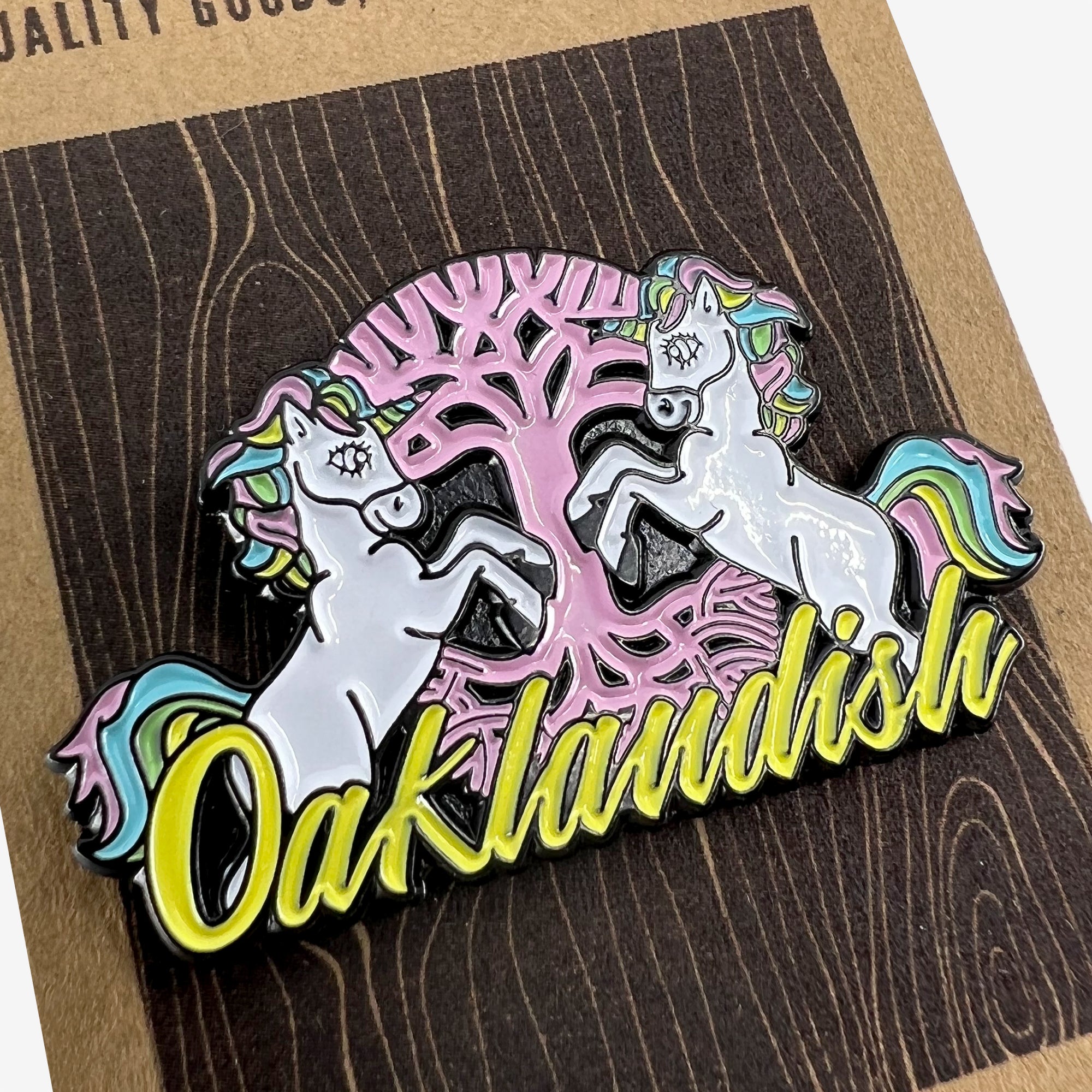 Close-up of enamel lapel pin with yellow Oaklandish wordmark, pink tree logo, and two white unicorns.