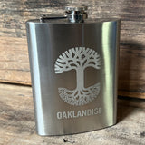 Silver 8 oz hip flask with screw top lid, gold Oaklandish tree logo, and wordmark on a wooden shelf.