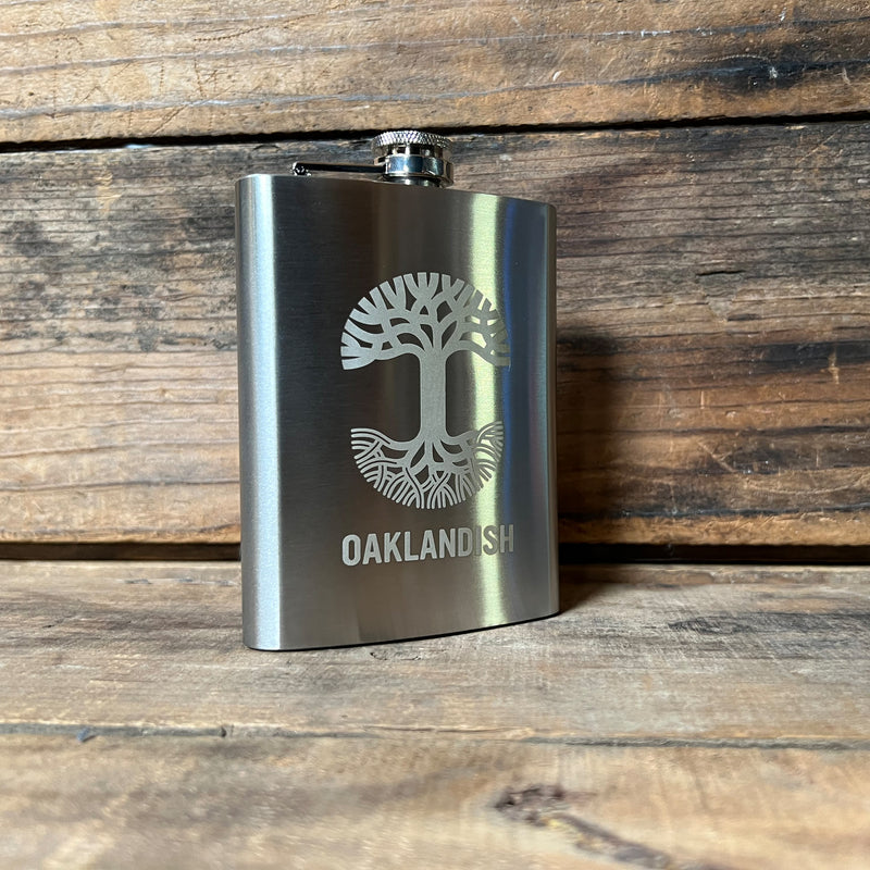 Silver 8 oz hip flask with screw top lid, gold Oaklandish tree logo, and wordmark on a wooden shelf.
