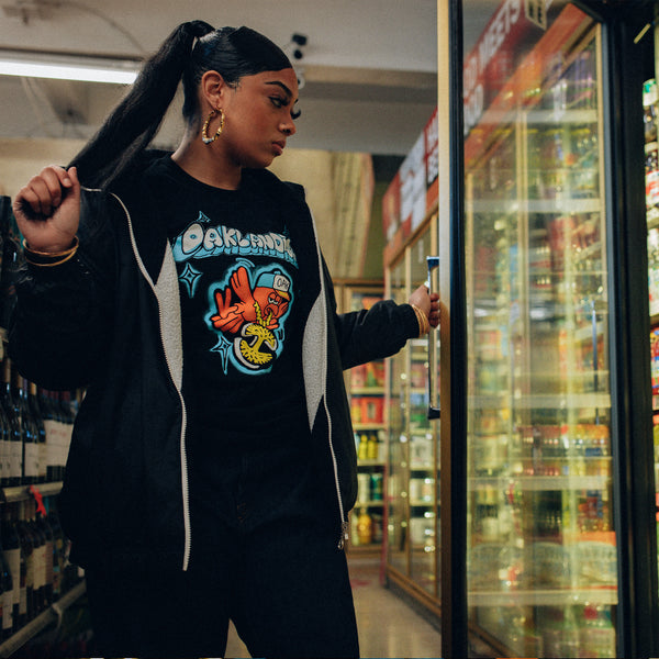 Female model wearing black t-shirt with a graphic image of a flying cartoon bird wearing an Oaklandish logo necklace with Oaklandish bubble wordmark above. while shopping in store.