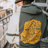Close up of a man wearing an army green premium Oaklandish pullover hoodie with a gold GATS mask silhouette on the back.