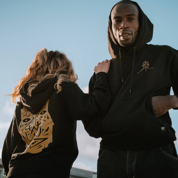 A man and a woman are standing outdoors, wearing black premium hoodies with gold GATS mask silhouette on the back and front left chest.