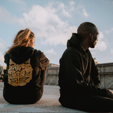 A man and a woman are sitting outdoors, wearing black premium hoodies with gold GATS mask silhouette on the back.