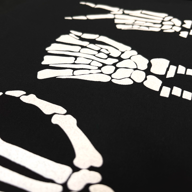 Detailed close-up of skeletal bone hands signing 'OAK' in white puff ink on a black t-shirt.