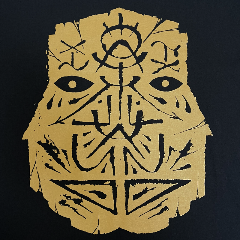 Detailed close-up of an oversized gold GATS mask silhouette on the chest of a black t-shirt.