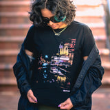 Female model wearing  black cotton t-shirt with an image of a rainy night reflecting street lights and The Grand Laker Theatre in Oakland.