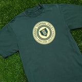 Green t-shirt with yellow Howard Terminal Field, Oakland California logo, and wordmark on the chest lying on the grass.