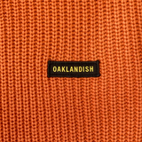 Detailed close-up of embroidered Oaklandish wordmark woven label patch on an autumn orange heavy-knit sweater.