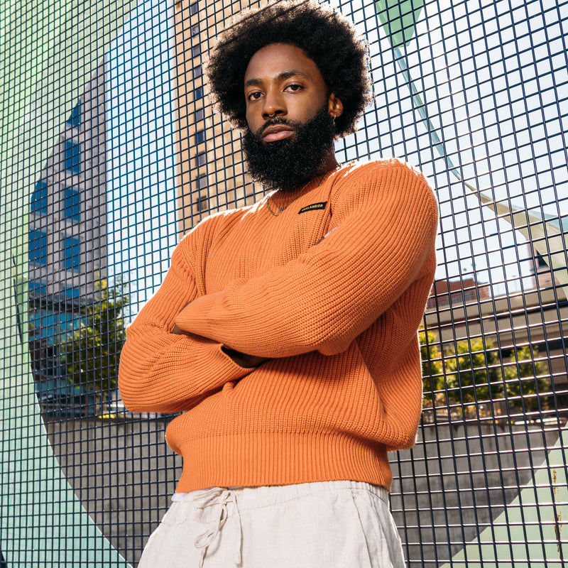 A man standing outside, from the waist up, in an autumn orange knit sweater with embroidered Oaklandish wordmark label on the front left chest.