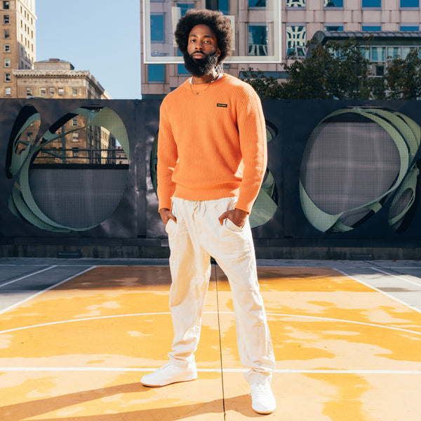 A man standing outside in a heavy cotton autumn orange knit sweater with embroidered Oaklandish wordmark label on the front left chest.
