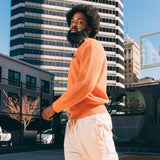 A man walking outside, from the waist up, in an autumn orange knit sweater with embroidered Oaklandish wordmark label on the front left chest.