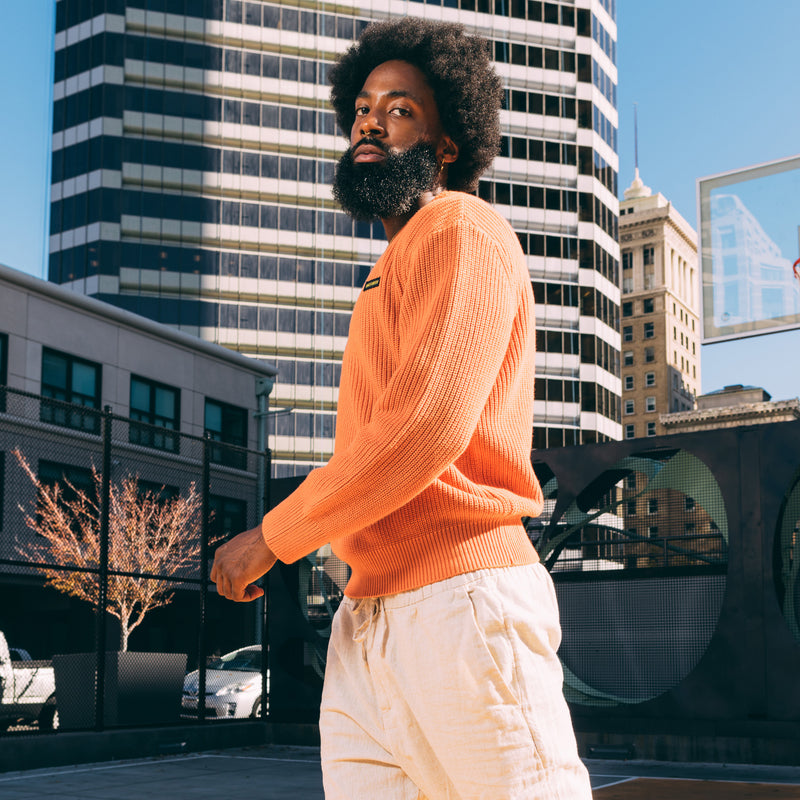 A man walking outside, from the waist up, in an autumn orange knit sweater with embroidered Oaklandish wordmark label on the front left chest.
