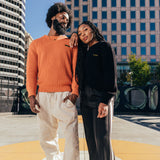 A man and a woman standing outdoors in Oakland in heavy-knit Oaklandish sweaters, with the man in autumn orange and the woman in black.