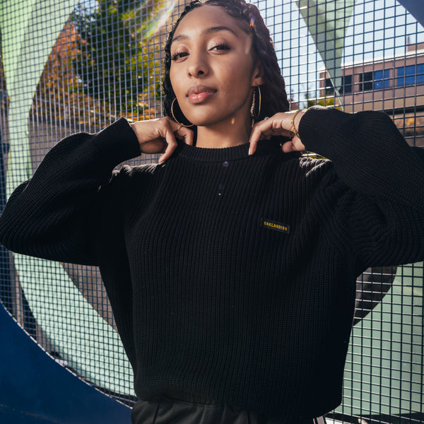 A woman outside, from the waist up, in a black knit sweater with embroidered Oaklandish wordmark label on the front left chest.