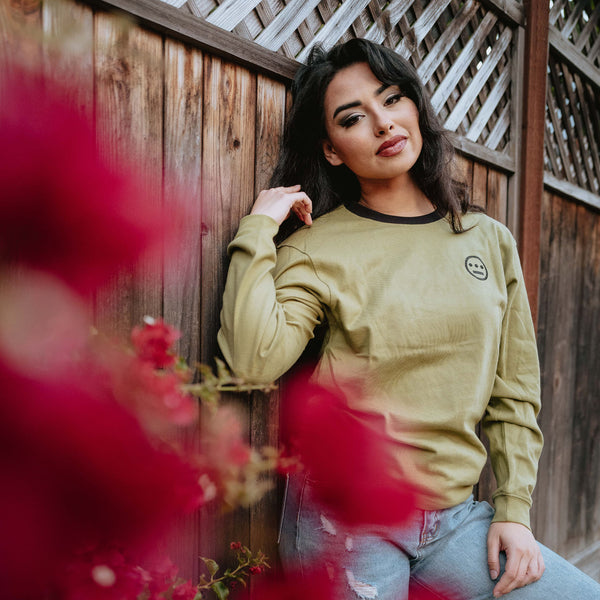 Army green long sleeve crew neck tee with black Hiero hip-hop logo on chest on woman outdoors leaning on a fence.