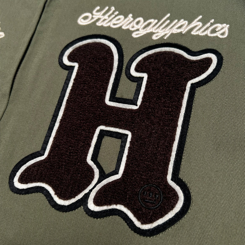 Detailed close-up of large chenille 'H' patch and cursive Hieroglyphics wordmark on left wear side chest of army green cadet jacket.