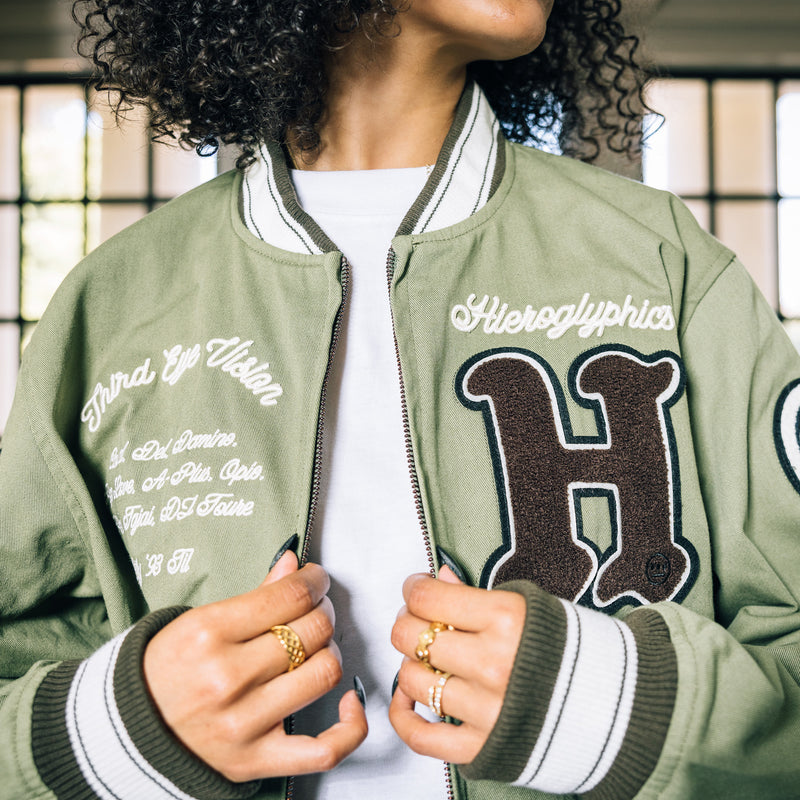 Close-up of a woman’s chest wearing an army green zip-up cadet jacket with large chenille Hiero H and cursive embroidered Hierogplyphics wordmark on left wear chest.