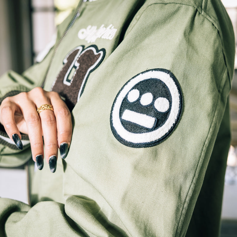 Woman wearing a army green cadet jacket with a detailed close up of chenille Hiero logo patch on left sleeve.