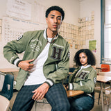 Man and woman in a high school classroom wearing army green zip-up cadet jackets with chenille Hiero Collective logo, wordmark patches on both sleeves, and left chest and third eye vision copy in cursive.