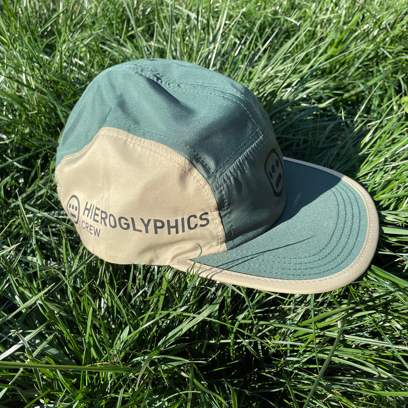 Side view of khaki side of a baseball cap with Hieroglypphics Crew logo and wordmark on the grass.