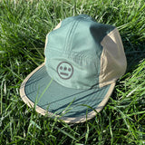 Camp style baseball cap with olive green crown and khaki sides with Hieroglyphics logo on crown sitting on grass.