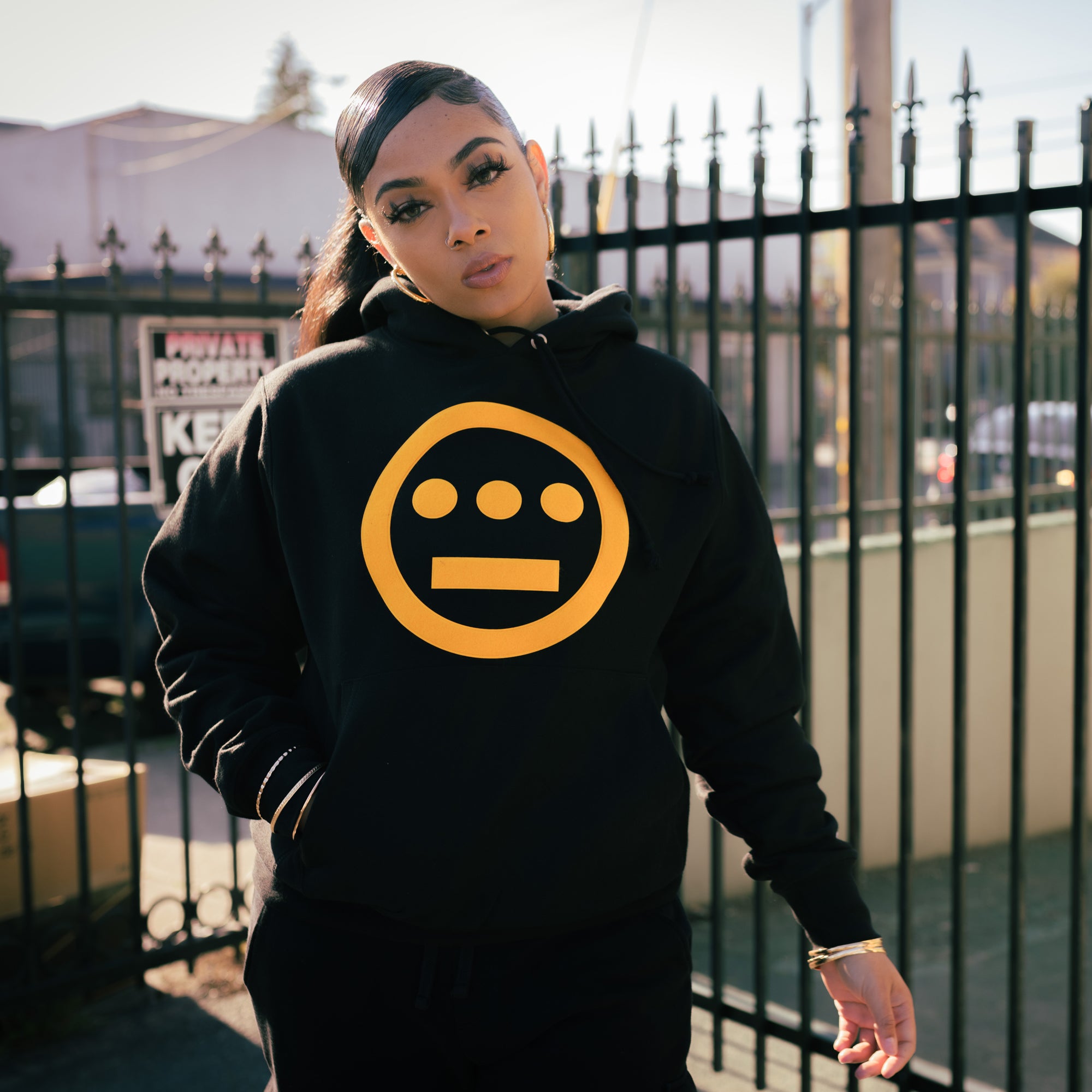 Female Model wearing champion x Hiero hoodie in black and yellow with one hand in front kangaroo pocket.