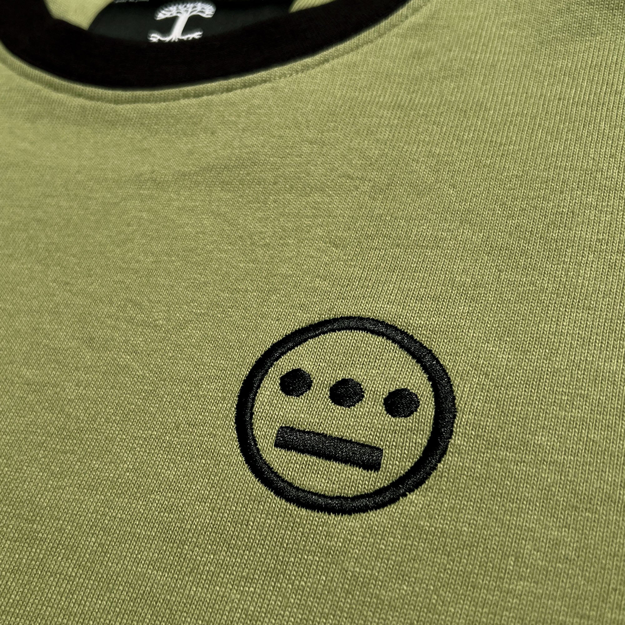 Detailed close-up of black embroidered Heiro hip-hop crew logo and black neck trim on an army green long-sleeve t-shirt. 