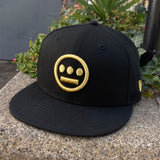 Front view of black New Era fitted cap with gold embroidered Hiero hip-hop logo on the crown sitting outdoors.
