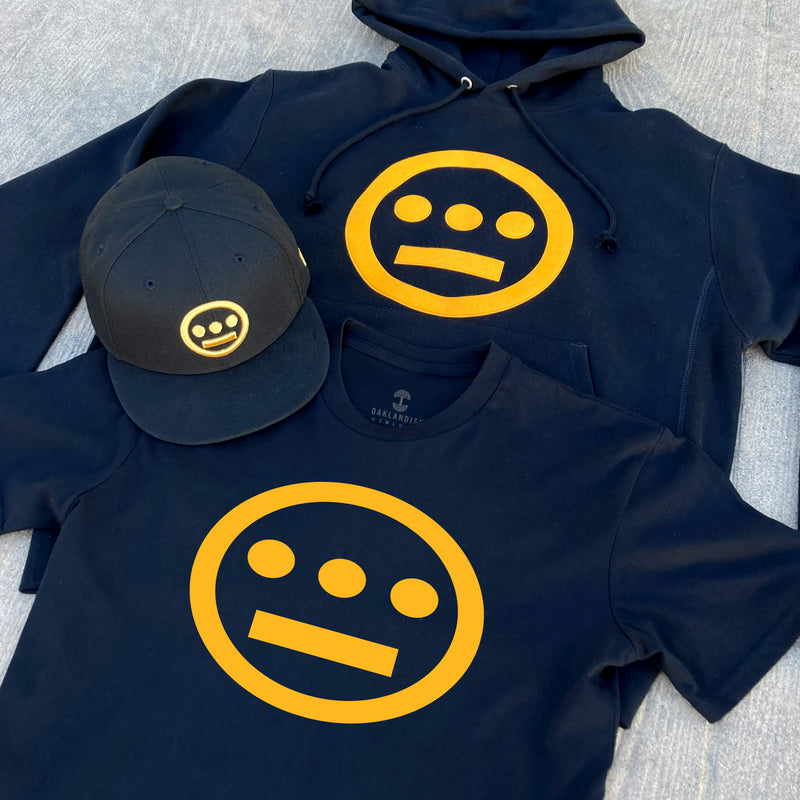 Black t-shirt, hoodie, and cap, each with a gold Hiero hip-hop logo on asphalt.
