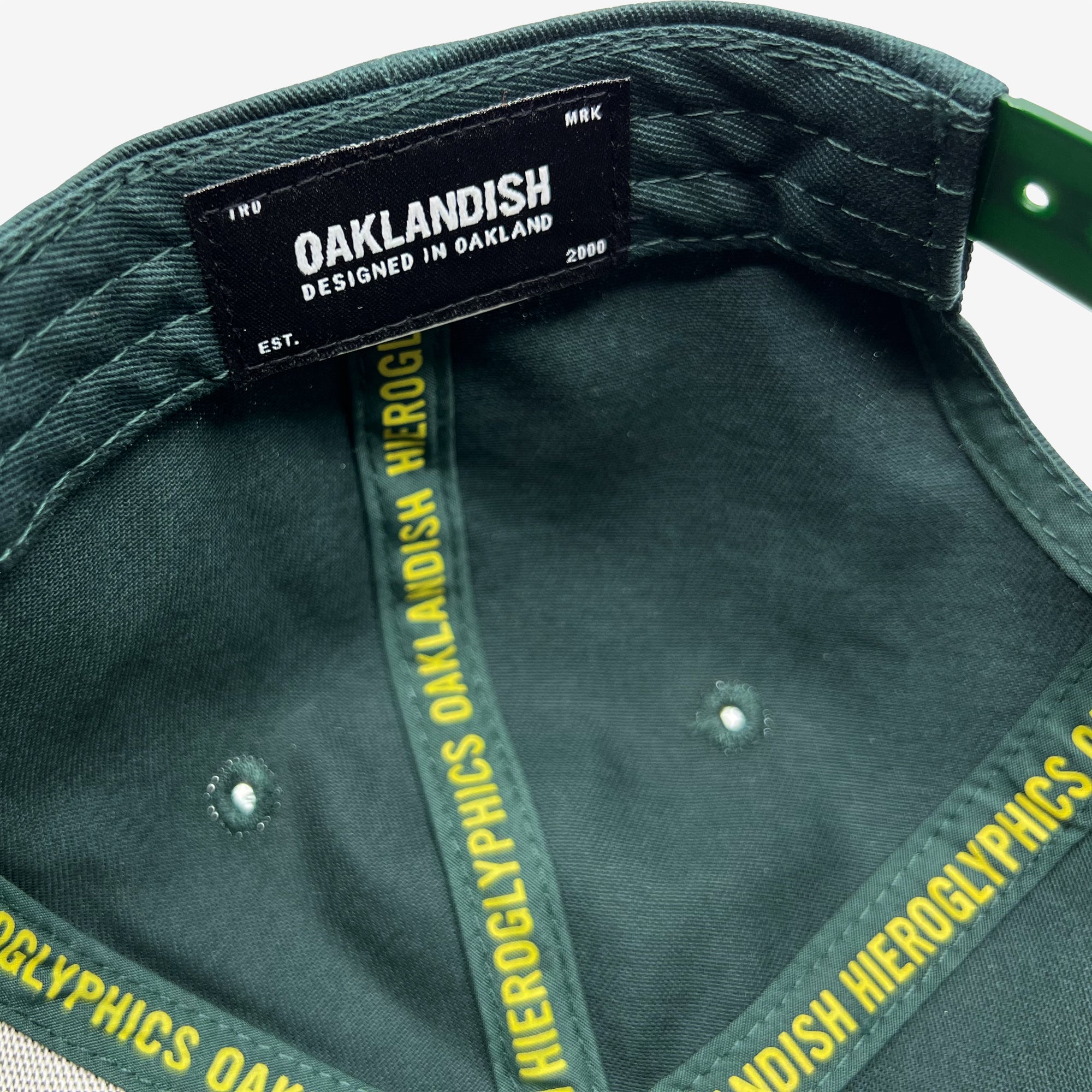 Close-up of the inside of a Hiero Logo Snapback cap by Hieroglyphics, featuring a black label sewn into it. The label reads "OAKLANDISH Designed in Oakland EST. 2000." The inner tape showcases repeated yellow text saying, "HIEROGLYPHICS OAKLANDISH HIEROGLYPHICS." A green adjustable strap is also visible.