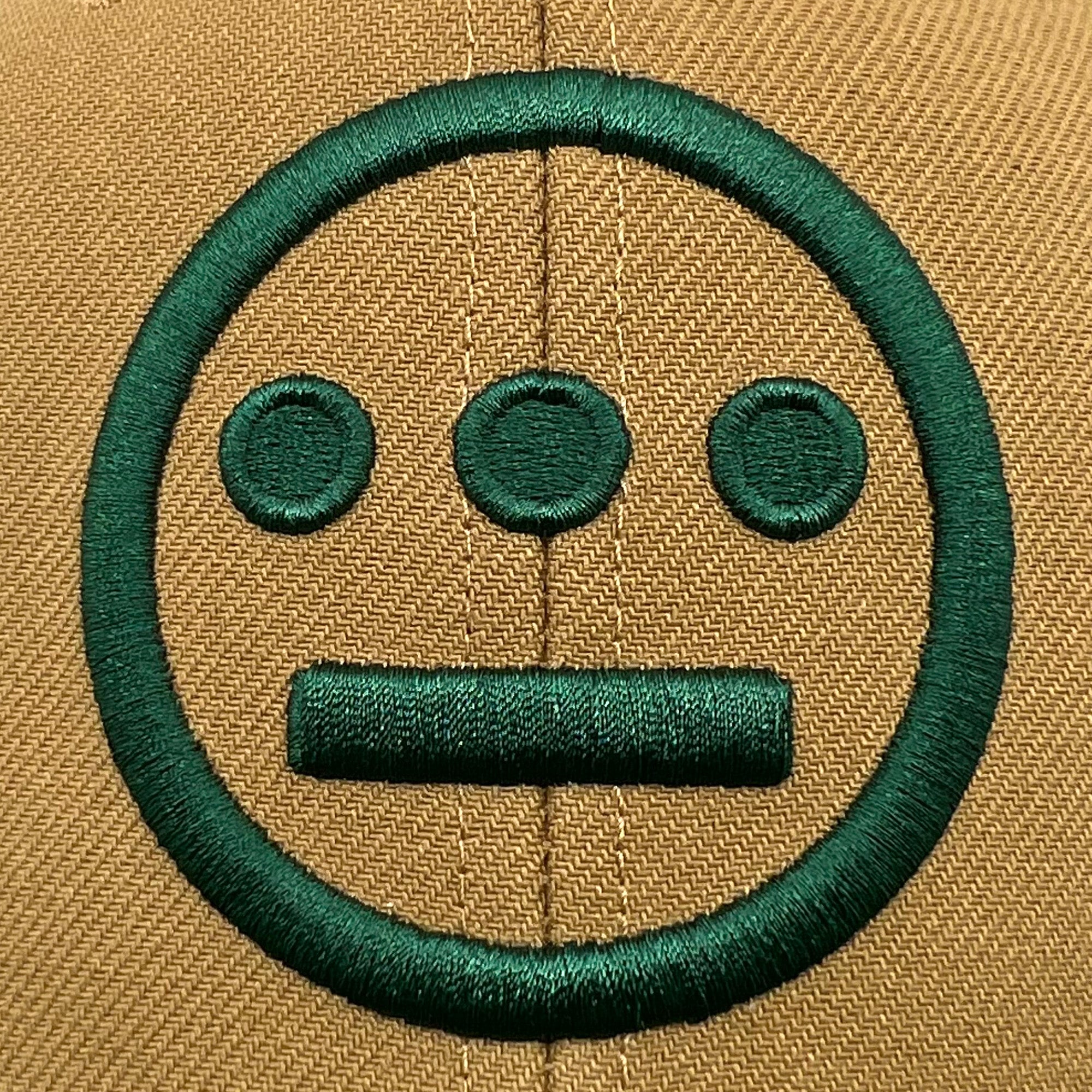 A beige fabric background features a prominent green embroidered design, reminiscent of the Hiero BNB Logo Snapback by Hieroglyphics. The design consists of a large circle encompassing two smaller circles positioned horizontally near the top and a horizontal bar shape underneath, resembling a face with a neutral expression.