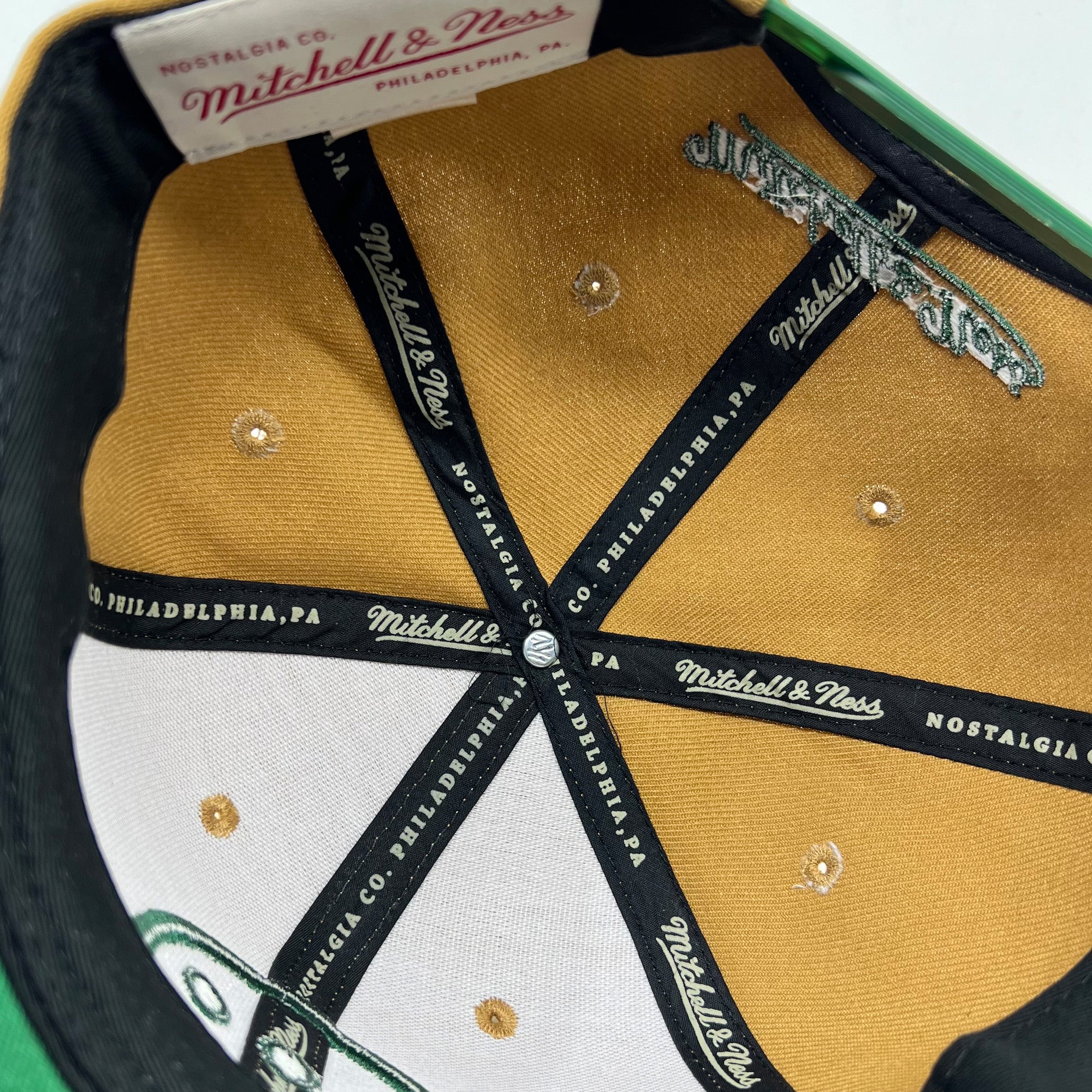 The inside of a Hieroglyphics Hiero BNB Logo Snapback hat in green is displayed, showcasing multiple black strips with "Mitchell & Ness" printed on them. The underbrim features embroidered details, and the inner label reads "Nostalgia Co. Mitchell & Ness Philadelphia, PA." The fabric and stitch details are prominently visible.